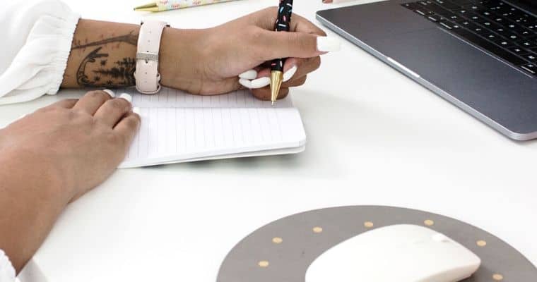 A person writing in a notebook next to a laptop and a mouse pad on a desk. The post is talking about how to write the best headlines for your blog.